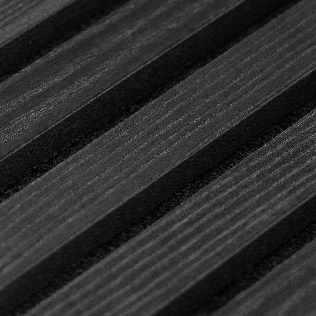 Close-up van een WoodUpp slatpaneel: smalle zwarte/antraciet houten latten met zichtbare nerf, matte afwerking en gelijkmatige diepe groeven ertussen. Diagonaal gefotografeerd, met hoog contrast tussen latten en schaduwen, benadrukt het strakke, moderne karakter van het WoodUpp lamellenpaneel, met nauwgezette afwerking en subtiele houttextuur zichtbaar.