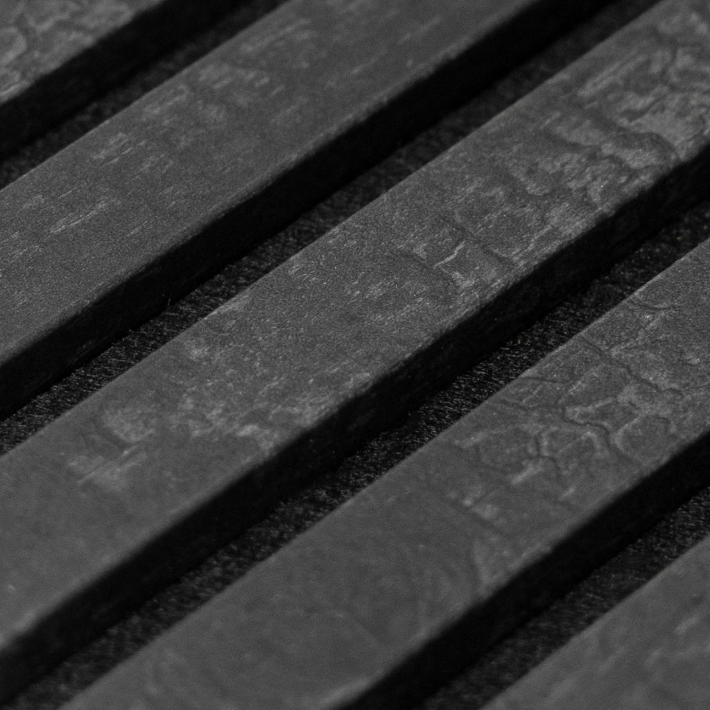 Close-up van WoodUpp zwarte, smalle houten latten die diagonaal over de foto liggen; vierkante, parallelle lamellen met matte, licht gegolfde houtnerf en subtiele marmerachtige textuur, geplaatst op een donkere, lichtkorrelige achtergrond — detailopname van WoodUpp lamellenpaneel. Met strakke randen, zichtbaar reliëf en hoogwaardige afwerking, ideaal voor moderne wandbekleding.