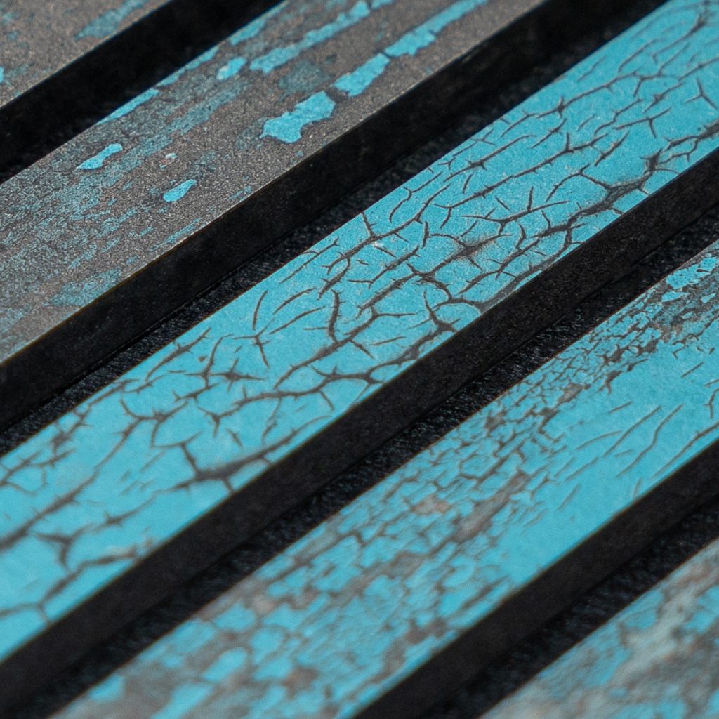 Close-up van WoodUpp houten strips met turquoise craquelé-lak: rechthoekige, donkergerande lamellen met gebarsten verfpatronen, spikkelige plekken en een matte, licht geruweerde textuur. De WoodUpp-producten tonen contrasterende zwarte tussenruimtes en variërende craquelé-patronen op elk smal paneel. Opname benadrukt de diagonale rangschikking en fijne structuurdetails.