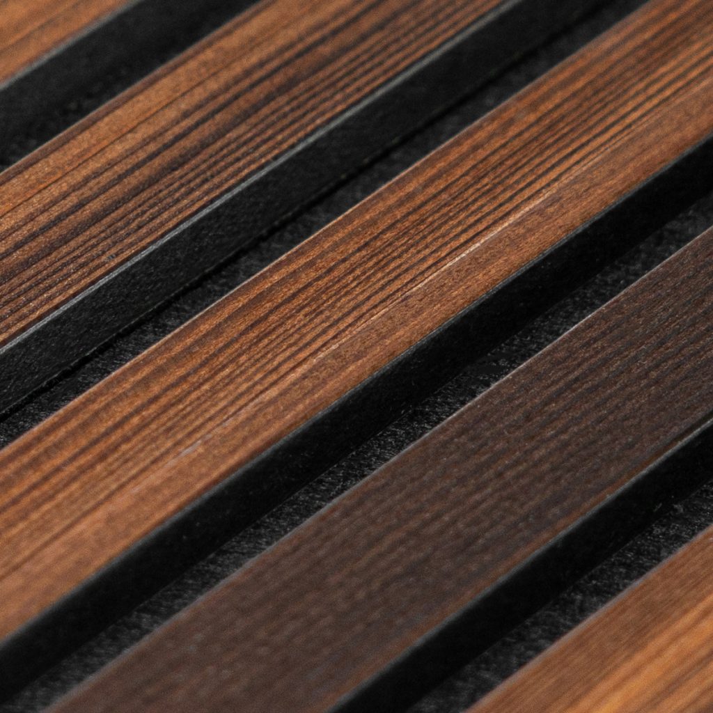 Close-up van een WoodUpp lamellenpaneel: parallelle, warmbruine houten latten met zichtbare nerf, afgewisseld door smalle zwarte tussenprofielen. Het beeld toont de fijne afwerking, contrast tussen natuurlijk hout en matte zwarte stroken, en het decoratieve, structurele karakter van WoodUpp lamellenproducten, duurzaam design en hoogwaardige materialen.