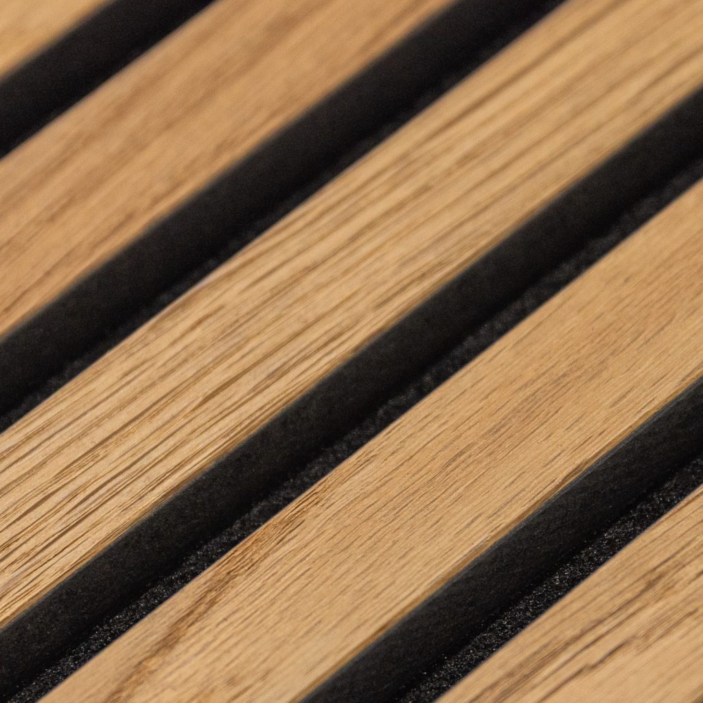 Close-up van WoodUpp houten lamellenpaneel: smalle, lichtbruine eikenhout-lamellen met zichtbare houtnerf, afgewisseld door diepe zwarte voegen gevuld met akoestische foam. Strak, diagonale compositie toont textuur en contrast tussen warme houttinten en matte zwarte dempende strips van WoodUpp, nauwkeurig afgewerkt voor wand- of plafondbekleding.
