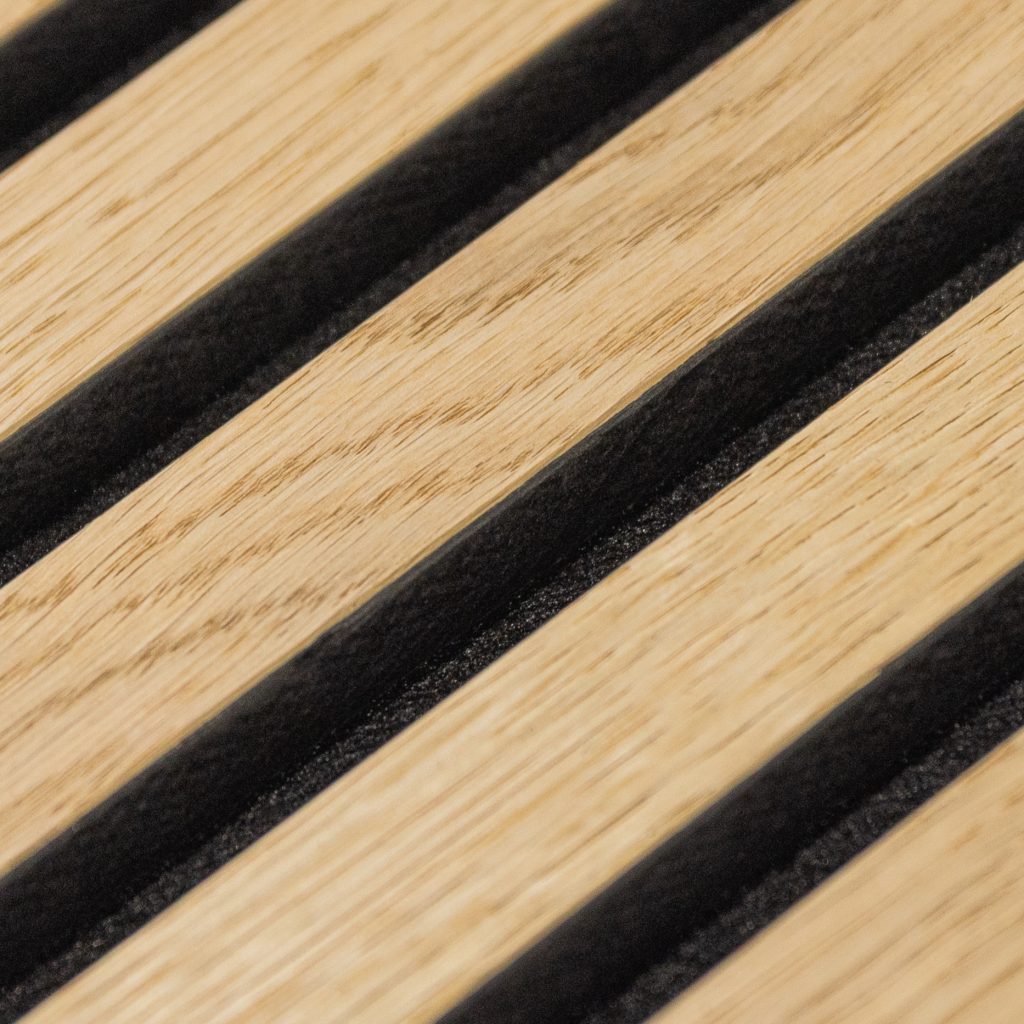 Close-up van een WoodUpp eiken lattenpaneel: diagonale lichtgekleurde eiken lamellen met zichtbare nerven, nauwkeurig afgewerkt en afgewisseld door smalle zwarte, geluiddempende stroken als achterlaag. Detail toont de houttextuur, het sterke contrast en de matte zwarte vulling van een WoodUpp slatwall akoestisch paneel.