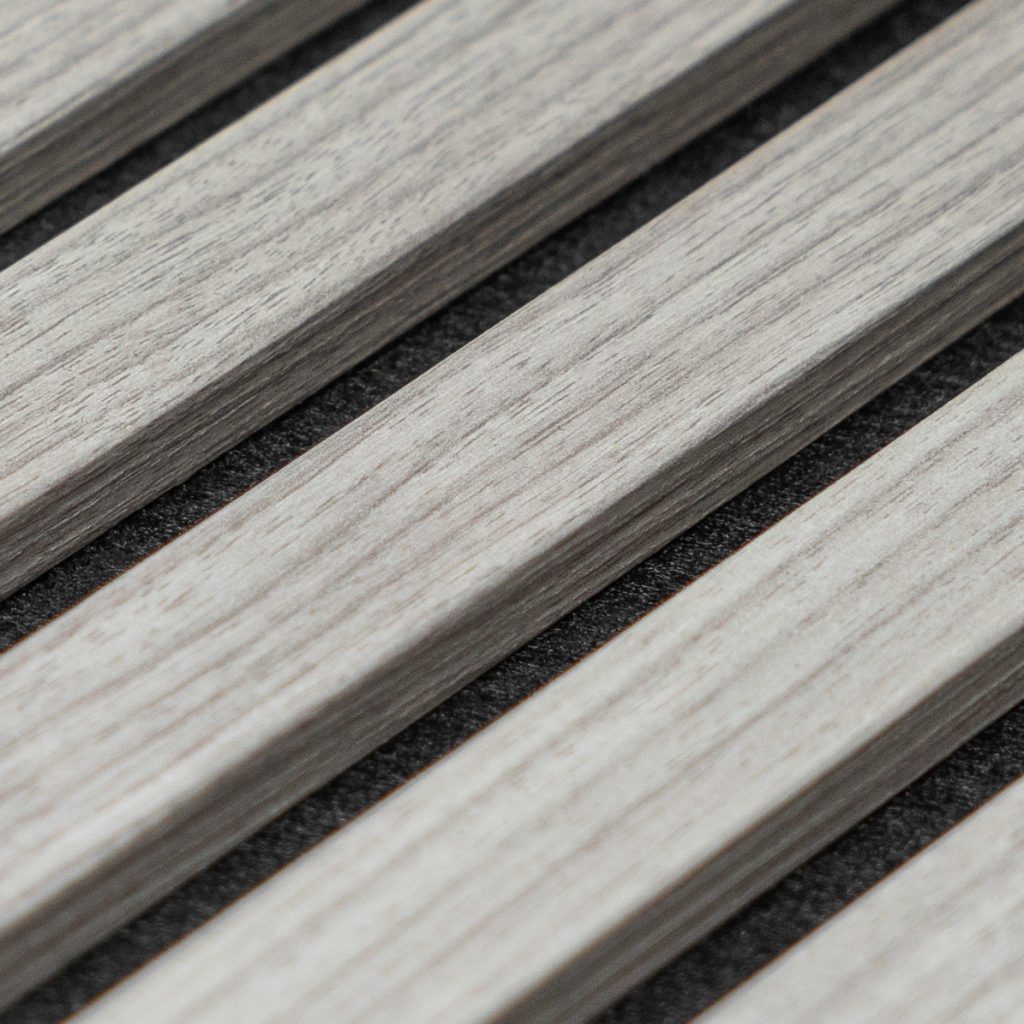 Close-up van WoodUpp houten lamellenpanelen: lichte, grijs-wash eikennerf lamellen strak naast elkaar met donkere, textiele akoestische onderlaag zichtbaar tussen de spouwen. Moderne slatpanelen voor muren of plafonds, textuur en fijne houttekening duidelijk in beeld. Detail toont afwerking, kleurvariatie en smalle voegen.