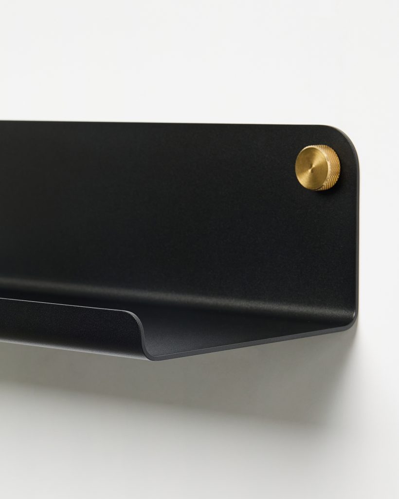 Zwarte metalen wandplank van WoodUpp met gebogen voorkant en subtiele opstaande rand, bevestigd aan de muur met een goudkleurige, geribbelde messing knop. Minimalistisch, mat oppervlak en afgeronde hoeken; geschikt als opbergplankje voor kleine objecten of display in modern interieur, stijlvol.