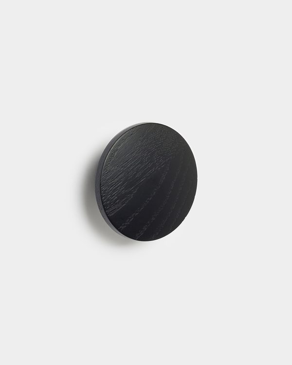 WoodUpp ronde zwarte houten wandhaak van massief eiken, met zichtbaar eikennerf en matte afwerking; een cirkelvormige knop gemonteerd op witte muur die een zachte schaduw werpt. Minimalistisch, compact design met dunne rand en natuurlijke houtstructuur, geschikt als kapstokhaak of decoratief accent.