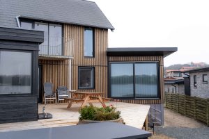 Modern huis met WoodUpp gevelbekleding: smalle verticale, naturel houten latten rondom grote zwarte kozijnen, en WoodUpp terrasplanken op het opgehoogde houten dek. Voorzijde toont picknickbank, stapelbare tuinstoelen en plantenbak; strakke, warme uitstraling door het verticale lattenpatroon en lichte dekplanken met uitzicht op water.