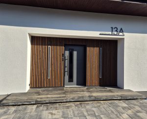 Voorgevel met modern zwarte voordeur omlijst door WoodUpp verticale houten lamellenpanelen aan beide zijden, twee slanke geïntegreerde wandlampen en WoodUpp houten terrasplanken/tegels op de opstap. De antracietgrijze deur heeft smalle glasstrook en lang zwart handvat. Huisnummer 13A zichtbaar rechtsboven op de witte stucgevel.