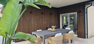 Overdekt terras met WoodUpp verticale, donkerbruine houten lamellen tegen de muur, geprofileerd met smalle zwarte tussenruimtes en zwarte afwerkingsprofielen. Schuifpui, grijze buiten-eettafel met rieten stoelen, groot bananenblad links en mandje met plant op tafel; drie kleine zwarte wandlampen verlichten de lamellen.