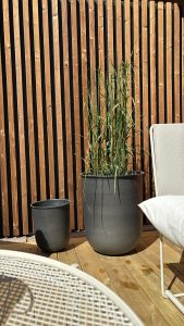 Zonovergoten beeld van een WoodUpp-achterwand met smalle verticale houten latten, zichtbaar houtnerf en warme amberkleur, en bijbehorende WoodUpp-terrasplanken. Voor staan twee antraciet plantenpotten (een grote met siergras), naast een wit lounge-stoeltje en deels zichtbaar rond metalen bijzettafeltje, met lichte zonneschaduwen op het hout.