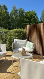 Zonnig terras met WoodUpp houten terrasplanken in natuurlijke kleur en een strak WoodUpp verticale lattenwand; lichte buitenlounge met zachte witgeklede kussens, ronde metalen salontafel, rieten lantaarn en een groen-wit deken op de stoel, omringd door groen geboomte onder blauwe lucht.