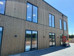WoodUpp-gevel van een twee verdiepingen tellend gebouw, bekleed met verticale houten latten van WoodUpp in natuurlijke houtkleur, met zwarte kozijnen rond grote ramen en glazen deuren. De slat-gevel toont ritme en schaduwwerking; een rode hoogwerker staat geparkeerd naast de ingang.