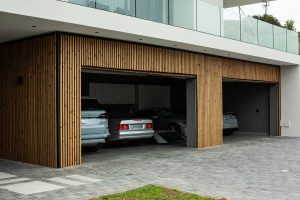 Moderne woning met drie open garagepoorten, bekleed met WoodUpp verticale houten lamellenpanelen in warme, natuurlijke tinten; slanke, gelijkmatig geplaatste latten vormen een sierlijke gevelbekleding en hoekafwerking rondom de garageopeningen, met meerdere geparkeerde auto's, bovenliggend glazen balkon en grijze oprit zichtbaar.
