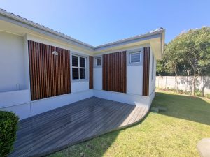 Hoek van een witte woning met WoodUpp houten lattenpanelen tegen de gevel (donker gebeitst, verticale smalle lamellen) rondom ramen, en een WoodUpp grijs terras van smalle planken composiet met afgeronde rand. Zonnige tuin met gazon en bomen op de achtergrond.