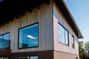 Hoek van een modern huis met WoodUpp verticale houten lattenpanelen als gevelbekleding op de bovenverdieping; strakke zwarte raamkozijnen en grote ramen, glazen balkonbalustrade, bakstenen plint en overstekend dak. De WoodUpp latten geven een slanke, ritmische, licht- en schaduwrijke uitstraling.