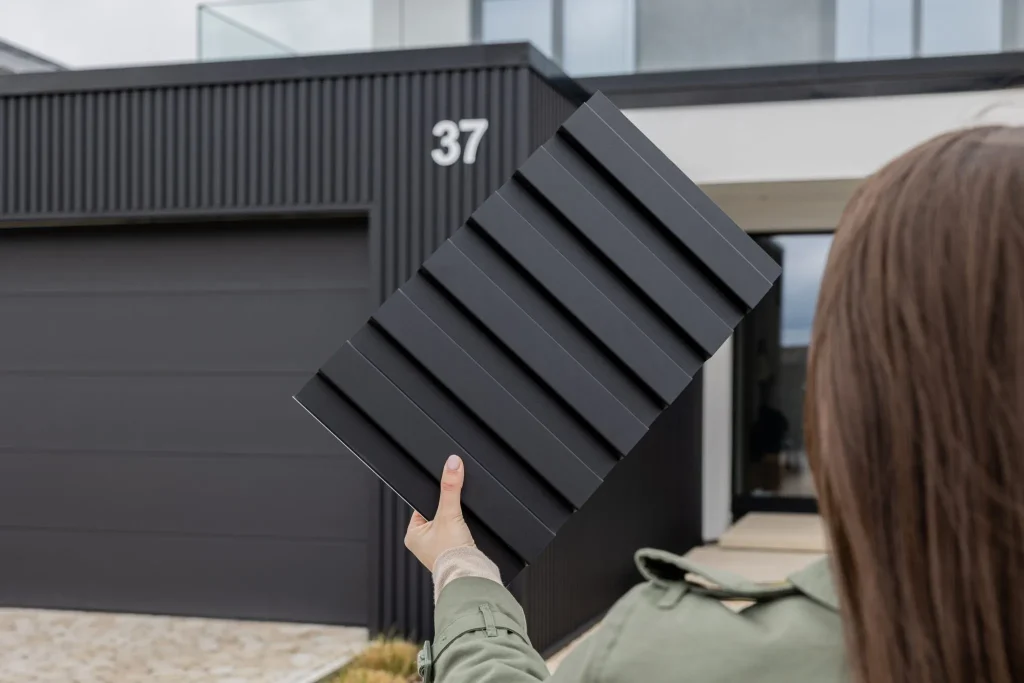 Hand houdt een WoodUpp monsterpaneel van zwart, geribbeld lamellenpaneel voor een moderne woning. Te zien zijn WoodUpp matte, donkergrijze gevelpanelen met diepe, gelijkmatige ribbels en dezelfde verticale bekleding op het huis en garage, met huisnummer 37 op de achtergrond en benadrukken een strakke, tijdloze uitstraling.