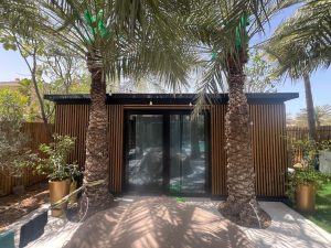 WoodUpp buitenruimte met moderne tuinunit bekleed met WoodUpp verticale houten lattenpanelen als gevelbekleding rondom zwarte glazen schuifdeuren. Voor de unit twee palmbomen en sierpotten, zonneschaduw en buitenlampjes zichtbaar; strakke latten bieden privacy en sfeervolle verlichting boven de deur.