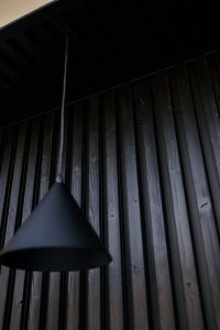 Zwarte WoodUpp verticale houten gevel- of wandlatten met donkere nerf en zichtbare schroefkoppen, gecombineerd met een matte zwarte conische WoodUpp-hanglamp aan een lange kabel, gefotografeerd van onderen tegen een donker plafond, met zichtbare textuur en natuurlijke knoesten — strakke, moderne houten afwerking en verlichting.