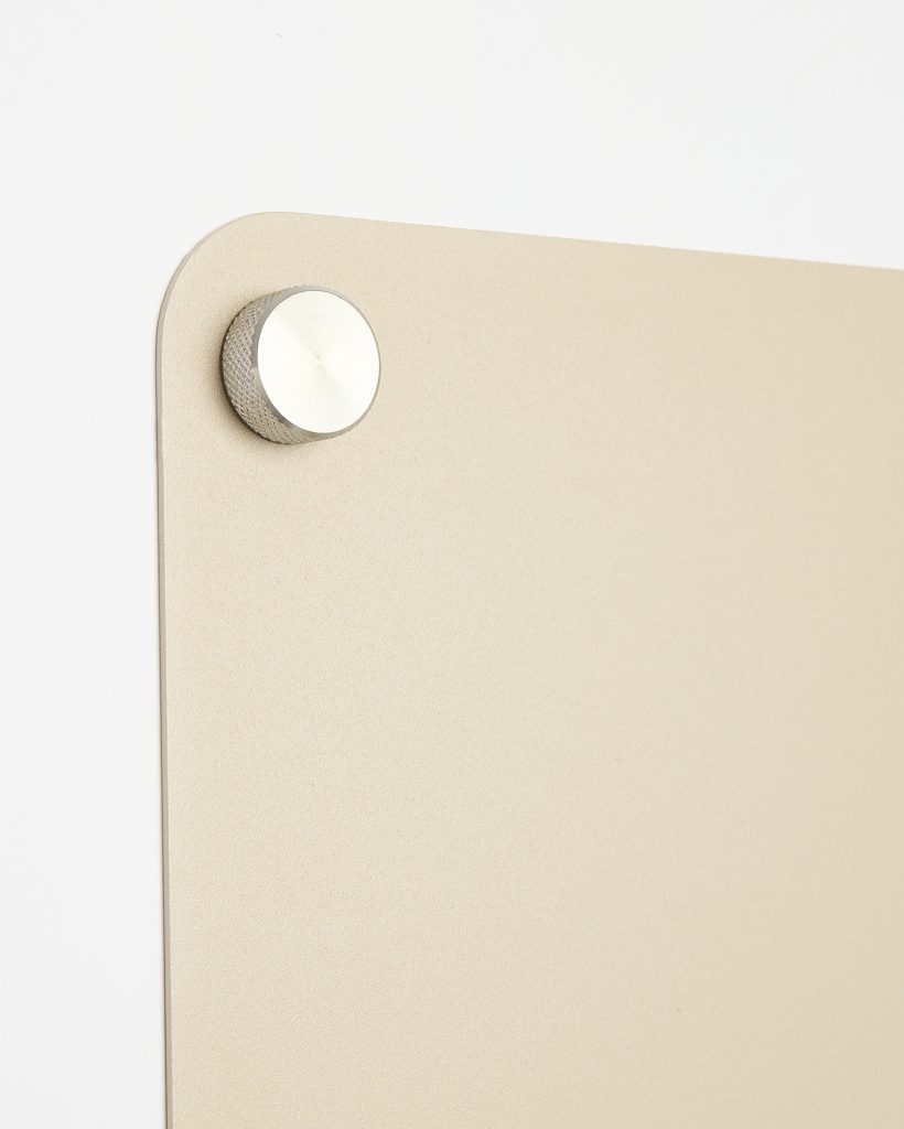 WoodUpp wandpaneel, groot rechthoekig en zandbeige met afgeronde hoeken en matte textuur, elegant en subtiel, zichtbaar in close-up. Paneel is bevestigd met een ronde zilverkleurige, geribbelde metalen montagestift/afstandhouder. Strak, minimalistisch WoodUpp-ontwerp dat het paneel van de witte muur plaatst.