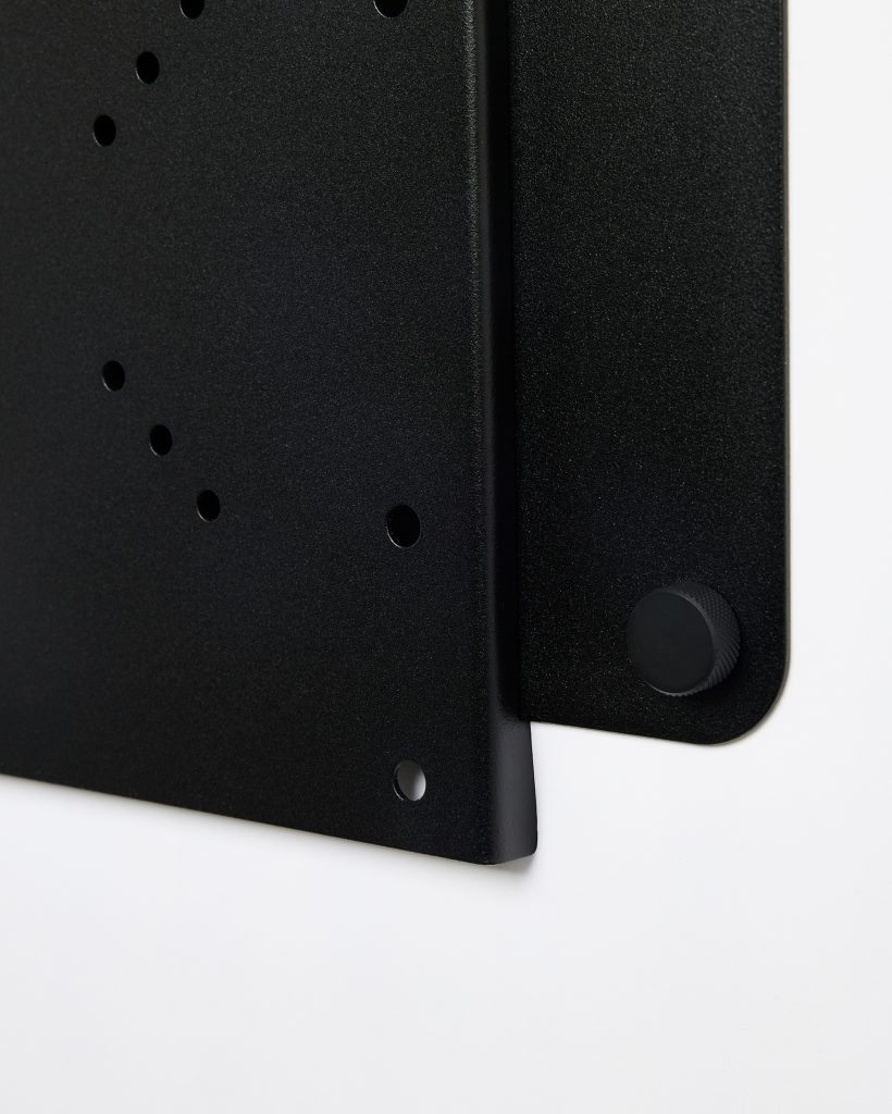 Close-up van WoodUpp metalen wandbeugels: twee matte zwarte, geperforeerde montageplaten met ronde bevestigingsgaten en afgeronde hoek, naast een geribbelde zwarte draaiknop/spacer. Strak gepoedercoate finish en overlap van platen tonen het minimalistische, functionele ontwerp van WoodUpp wand- en plankbevestigingen met stevige constructie.