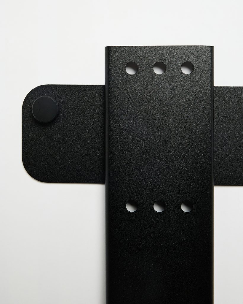 WoodUpp zwarte metalen wandbevestiging: twee overlappende matte plaatprofielen met afgeronde hoeken, een horizontale plaat met zichtbare ronde knop en een verticale strip met twee rijen van drie ronde bevestigingsgaten. Close-up toont mat zwarte poedercoating, strakke randen en witte achtergrond minimalistisch.