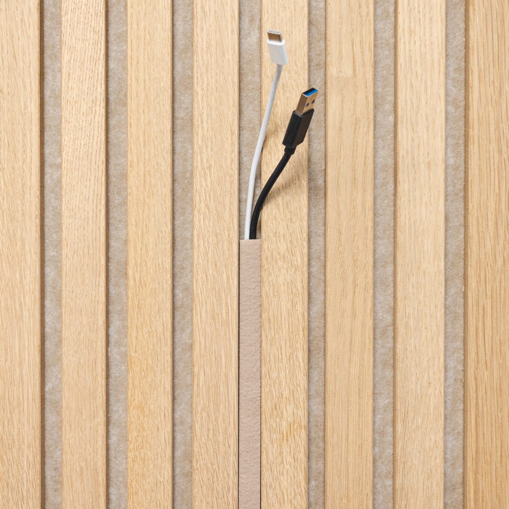 WoodUpp eiken lattenpaneel met smalle verticale kabeldoorvoer; lichte eikenhouten latten geplaatst boven grijze akoestische viltachterwand. In de opening steken twee kabels (witte USB‑C en zwarte USB‑A). Natuurkleurige matte finish, nauwkeurig gefreesde sleuf, discreet en geschikt voor woningen en interieurs.