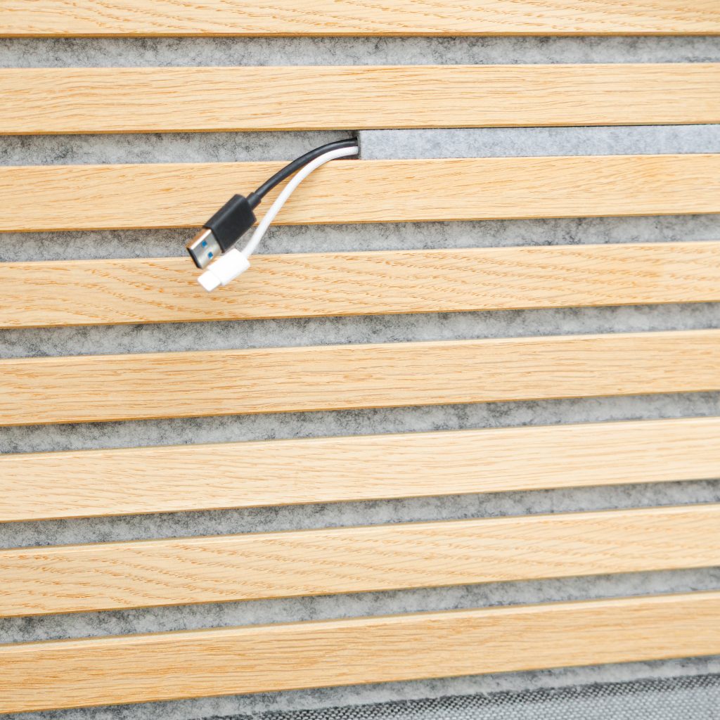 WoodUpp houten lattenpaneel van lichte eikenlook met zichtbare nerf en grijze akoestische viltachtergrond; smalle uitsparing in één lat toont een doorgang waar een zwarte USB-A-kabel en een witte magnetische oplaadkabel uitsteken, functionele afwerking voor kabelbeheer en geluidsabsorptie en strak modern interieuraccent.