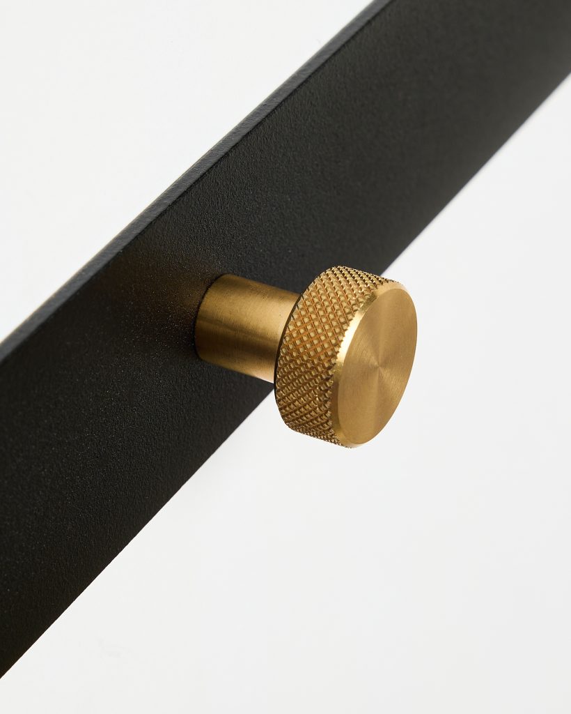 Close-up van twee WoodUpp-producten: een matte zwarte metalen plaat en een goudkleurige messing knop/haak. De knop heeft een cilindrische schacht, een grof geribbeld (knurled) rand en een glad rond voorvlak, met scherpe details, contrast tegen de witte achtergrond en subtiele schaduwwerking.