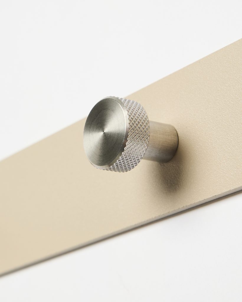 Close-up van een WoodUpp-product: een geborstelde metalen knop met geribbeld knurling, bevestigd op een smalle zandkleurige beige metalen wandplaat. De cilindrische knop heeft een ronde, licht conische kop en korte stift, met fijne draaisporen zichtbaar en een matte metallic afwerking die een moderne, elegante uitstraling geeft.