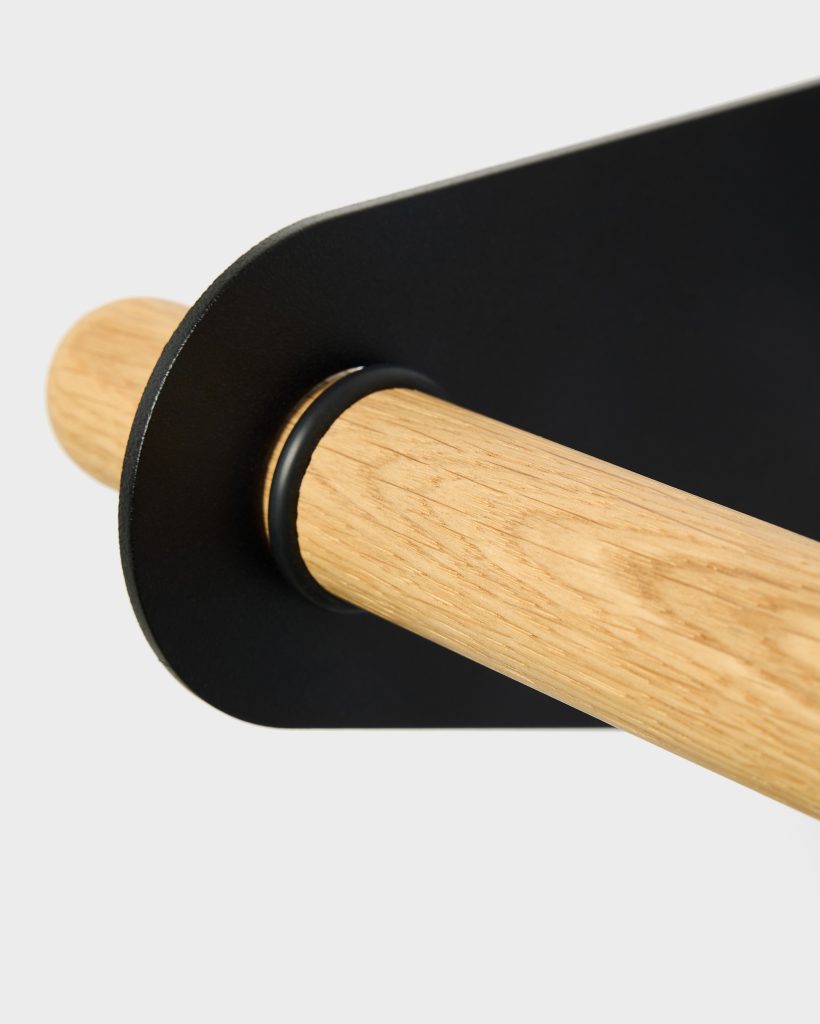Close-up van een WoodUpp wandrek: een eikenhouten stok met zichtbare nerf gesteund door een matte zwarte metalen beugel met afgeronde hoek en zwarte rubber ring rond de pen, benadrukt het minimalistische Scandinavische ontwerp en de hoogwaardige afwerking, voor gebruik als handdoekhouder of kledingstang.