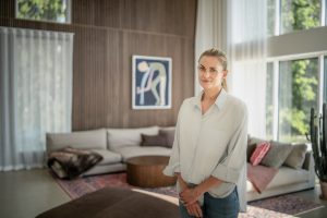 Vrouw staat in modern woonkamertje met WoodUpp verticale houten lamellenwand en een rond WoodUpp houten salontafeltje voor een lichte hoekbank; de lamellen tonen warme houttint en ribbelstructuur, zachte kussens, kleurrijk vloerkleed, grote raampartij en een hoge cactus, vrouw in lichtblouse glimlacht rustig.