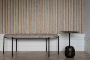 Foto van een rustige interieurhoek met WoodUpp houten lamellenpanelen tegen de muur: smalle verticale lichte lamellen met grijze tussenlaag, gecombineerd met een gestoffeerd bankje op zwarte metalen poten en een rond bijzettafeltje met dun zwart blad en donkere marmeren voet.