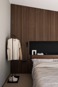 WoodUpp lamellenwand van donker walnoothout achter het bed, met bijpassend WoodUpp houten hoofdeinde en smalle nachtplank; minimalistische zwarte metalen kledingstandaard met hangende beige T‑shirts en witte sneakers op de voetplaat; bed met lichte dekens, strakke moderne uitstraling en warme tinten.