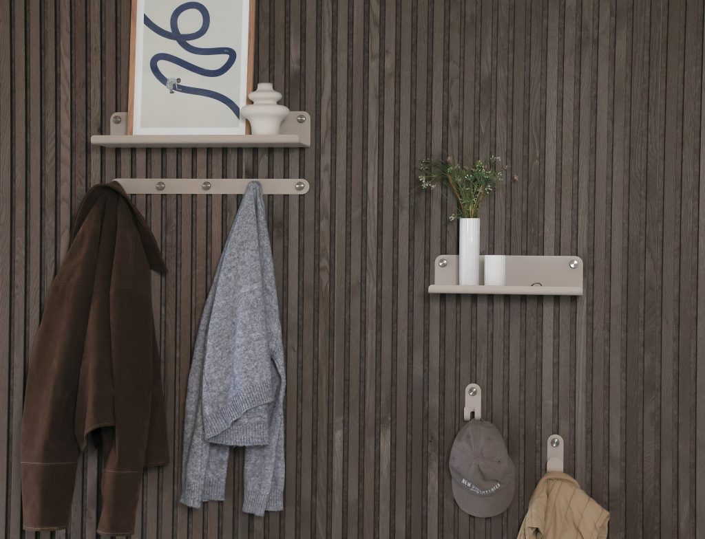 Donkere houten lambrisering met WoodUpp beige wandplanken en haken: boven links een WoodUpp plankje met lijst en keramisch vaasje, daaronder een WoodUpp kledingrail met metalen knopjes waaraan een jas en trui hangen. Rechts een WoodUpp plank met witte vazen en twee losse haken met pet en jas.