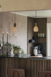 WoodUpp: donkere verticale lamellenpanelen bekleden zowel de achterwand als het front van het keukeneiland; zichtbaar zijn WoodUpp lamellenpanelen en lamellenbekleding. Voorgrond toont hanglampen, kaarsen, een groene plantenpot en een koffiezetapparaat op het aanrecht, in een warme, moderne keukensetting.
