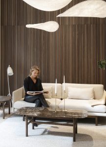 Vrouw leest op een crèmebank voor een WoodUpp houten lattenwand; op de vloer een WoodUpp donkerhouten salontafel met marmeren blad en afgeronde hoeken, plus een bijpassende bijzettafel. Bank met zachte kussens, twee slanke kaarsen en drie organische hanglampen completeren de warme interieursetting.