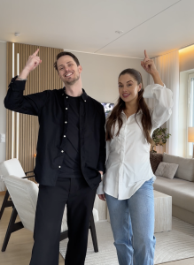 Een lachend koppel wijst omhoog in een moderne woonkamer. Op de achtergrond is een WoodUpp akoestisch wandpaneel te zien. Dit paneel heeft eikenhouten latten op een zwarte viltachtergrond en is voorzien van geïntegreerde verticale LED-stripverlichting tussen de latten.