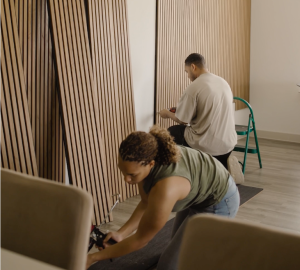 Een man en een vrouw zijn bezig met het installeren van WoodUpp akupanelen in een kamer. De vrouw brengt lijm aan op een paneel op de grond, terwijl de man een paneel aan de muur bevestigt. De wandpanelen zijn van licht eikenhout met een zwarte vilten achterkant.