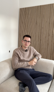 Een jongeman met een bril en een bruine trui zit comfortabel op een lichte bank. Achter hem is de muur bekleed met een stijlvol WoodUpp akoestisch paneel. Dit paneel kenmerkt zich door verticale eikenhouten latten op een zwarte vilten achtergrond, wat een warme en moderne uitstraling geeft.