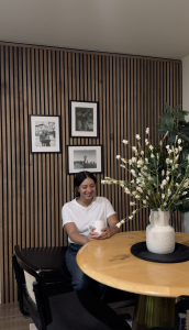 Een vrouw geniet van een kopje koffie aan een ronde tafel, omringd door een sfeervolle wand met WoodUpp Akupanelen. Deze houten lattenwand in een warme walnootkleur op zwart vilt creëert een gezellige en stijlvolle achtergrond voor het interieur.