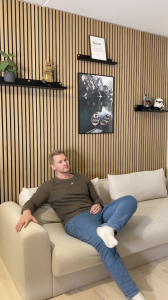 Een man ontspant op een beige bank, leunend tegen een muur volledig bekleed met WoodUpp Akupanel. Deze akoestische panelen hebben natuurlijk eikenhouten latten op een zwarte vilten achtergrond. Er zijn ook bijpassende zwarte WoodUpp planken gemonteerd, waarop decoraties zoals lijsten en Lego staan.