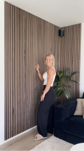 Een lachende vrouw poseert in een hoek die volledig is bekleed met stijlvolle WoodUpp Akupanelen. Deze akoestische houten lattenwand in een warme walnootkleur loopt door op twee muren in de moderne woonkamer, naast een grote plant en een donkere bank.