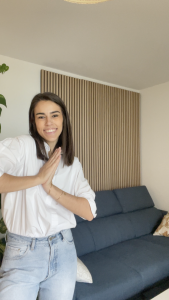 Een lachende vrouw in een wit overhemd staat voor een verticaal WoodUpp akoestisch wandpaneel met een lichte eikenhouten afwerking. Het houten lattenpaneel van WoodUpp is gemonteerd op de muur achter een blauwe bank in een lichte woonkamer.