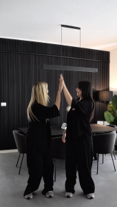 Twee vrouwen in zwarte kleding geven een high-five in een moderne eetkamer. De volledige achterwand is bekleed met zwarte WoodUpp Akupanel akoestische panelen, gekenmerkt door verticale zwarte houten latten op een donker vilt. Dit zorgt voor een stijlvol en geluiddempend effect in de ruimte.