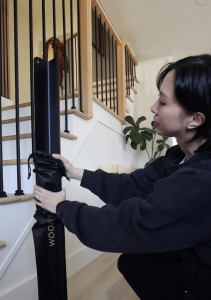 Een vrouw knielt op een houten vloer en houdt een lang WoodUpp product vast, verpakt in een zwarte beschermhoes met het gouden WoodUpp logo erop. Het object lijkt een zwarte aluminium afwerklijst of hoekprofiel te zijn, gebruikt voor de installatie van akoestische panelen, gesitueerd naast een trap.