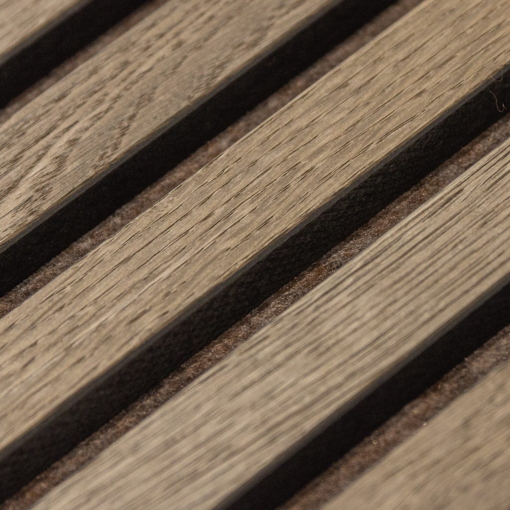 Close-up van WoodUpp houten lattenpaneel: schuine, smalle eikenhouten latten met zichtbare houtnerf en lichtgrijsbruine kleur, geplaatst boven donkere vilten tussenlagen voor akoestische demping. Detail toont de textuur en de smalle openingen tussen latten, geschikt voor moderne geluidsabsorberende wand- of plafondafwerking.