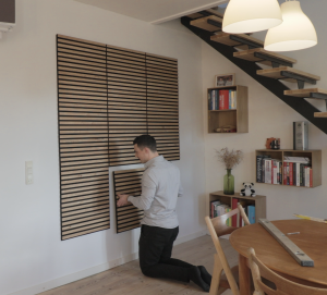 Een man is bezig met het installeren van vierkante WoodUpp Akupanelen aan een witte muur. Deze akoestische panelen hebben licht eiken houten latten op een zwarte viltachtergrond. Hij knielt op de vloer en plaatst zorgvuldig een van de panelen om de wandbekleding te voltooien in een sfeervolle woonkamer.