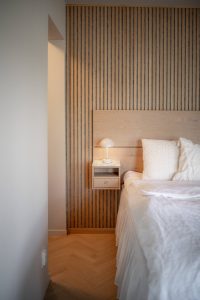 Minimalistische slaapkamerhoek met WoodUpp houten wandbekleding van smalle verticale latten, bijpassend WoodUpp massief houten hoofdbord en een zwevend WoodUpp nachtkastje in licht eiken met lade en licht marmeren blad; witte bedtextiel en een klein, sfeervol tafellampje completeren de warme, natuurlijke uitstraling