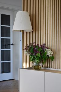 Sfeervol, rustig interieurbeeld van een WoodUpp eikenhouten lattenwandpanelen naast een wit dressoir met houten blad, een glazen vaas vol paarse en witte sering en een slanke staande lamp met witte geplooide kap; natuurlijke verticale latten creëren warme textuur en subtiele schaduwlijnen.