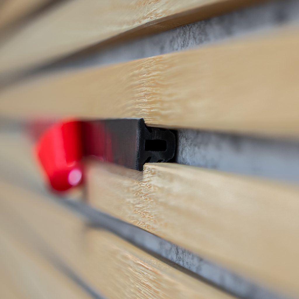 Close-up van WoodUpp lamellen: lichtgekleurde horizontale latten met zichtbare nerf en smalle tussenruimtes, gemonteerd op grijze ondergrond, nauwkeurig geplaatst, met ruwe houttextuur en onscherpe achtergrond, close-up perspectief. Duidelijk zichtbaar is het zwarte WoodUpp-montageprofiel met H-vormige sleuf en een rood vergrendelingsclipje tussen de latten.