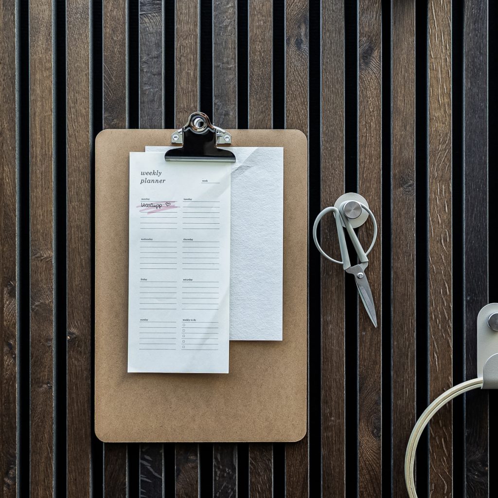 WoodUpp: bovenaanzicht van een donkere houten lattenwand met een WoodUpp clipboard (MDF) waarop een weekplanner-notitieblaadje is geklemd en een wit notitievel eronder. Rechts een WoodUpp magnetische haak met metalen schaar. Nette, minimalistische werkplekcompositie.